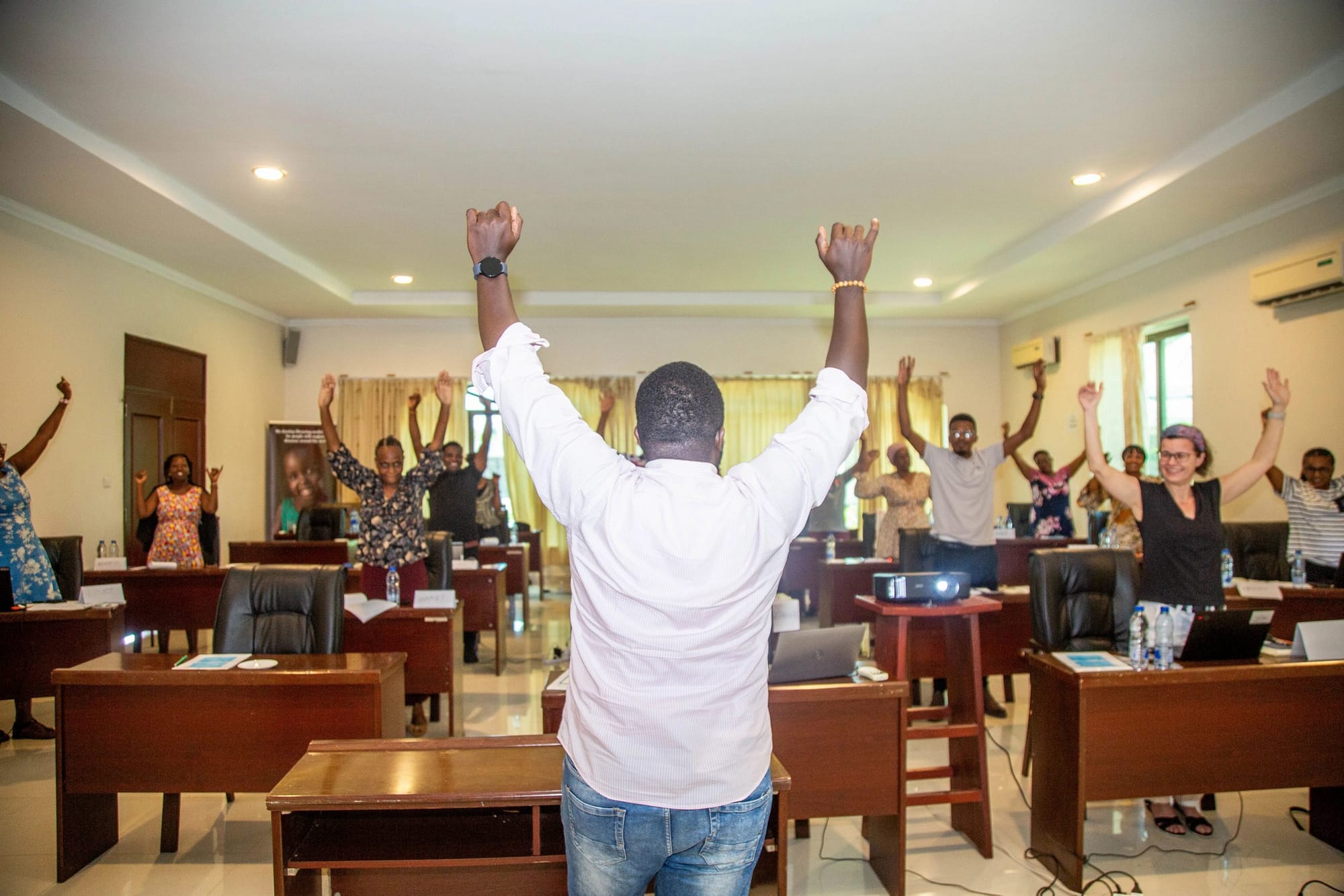 Man stands in front of a room of people with both hands raised